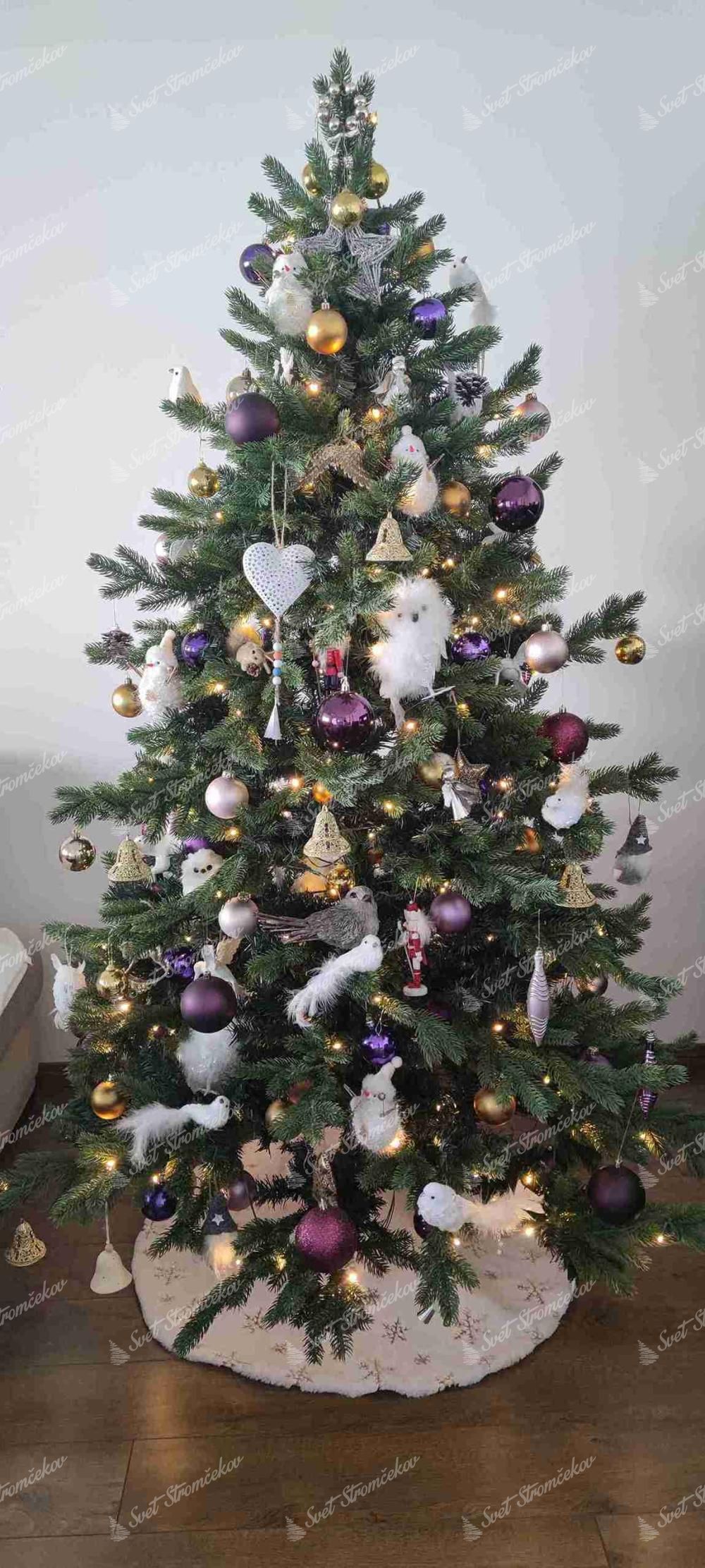 Umelý vianočný stromček 3D Smrek Exkluzívny 180cm LED300. Stromček so zeleným ihličím je ozdobený bielymi a fialovými ozdobami, vtáčikmi a sovami.