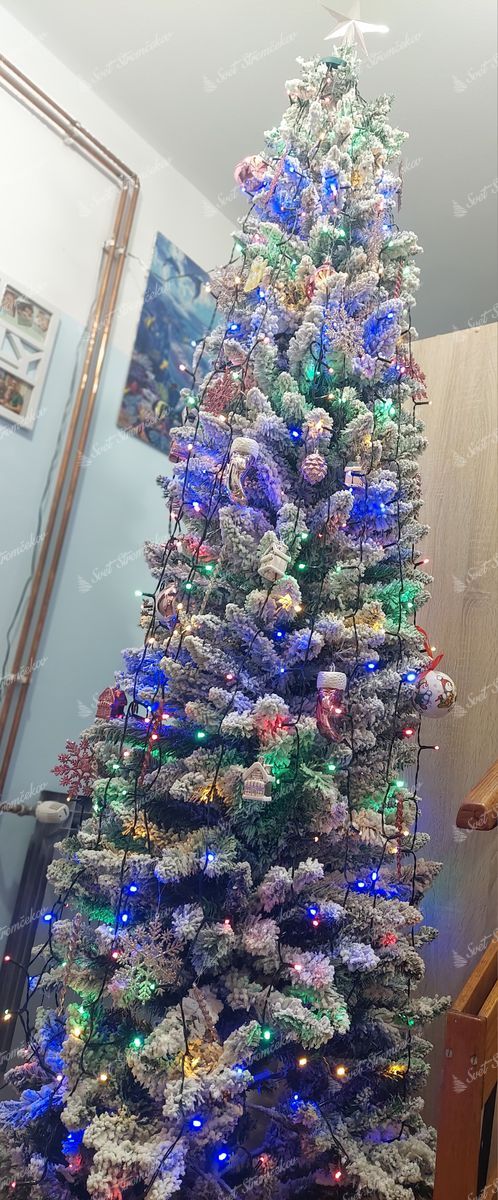 Umelý vianočný stromček Smrek Biely Štíhly 240cm, stromček je ozdobený malými ozdobami a farebným osvetlením.