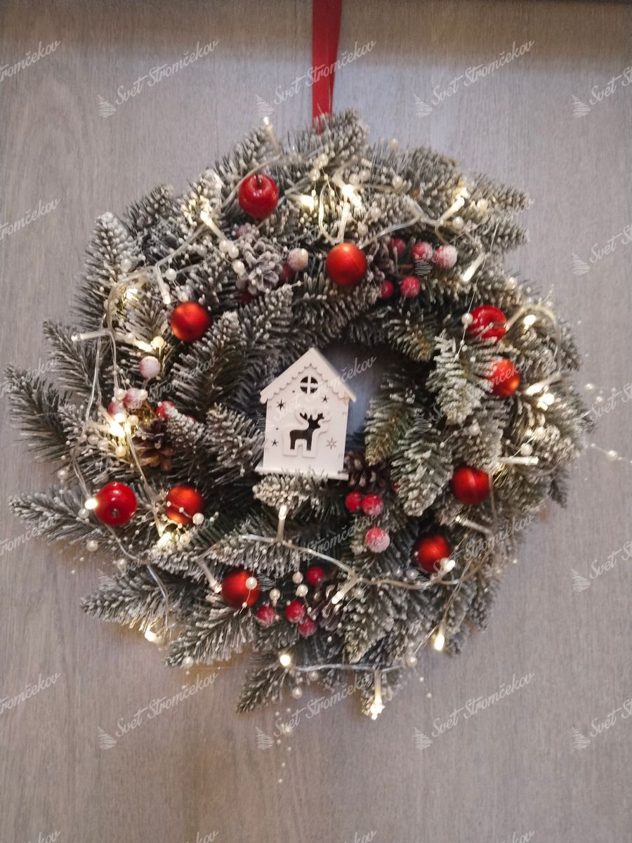 Vianočný veniec Smrek Kryštálový 50cm. Veniec má jemne zasnežené vetvičky, ozdobené šiškami a červenými plodmi a je ozdobený červenými ozdobami a drevenou dekoráciou domčeka.